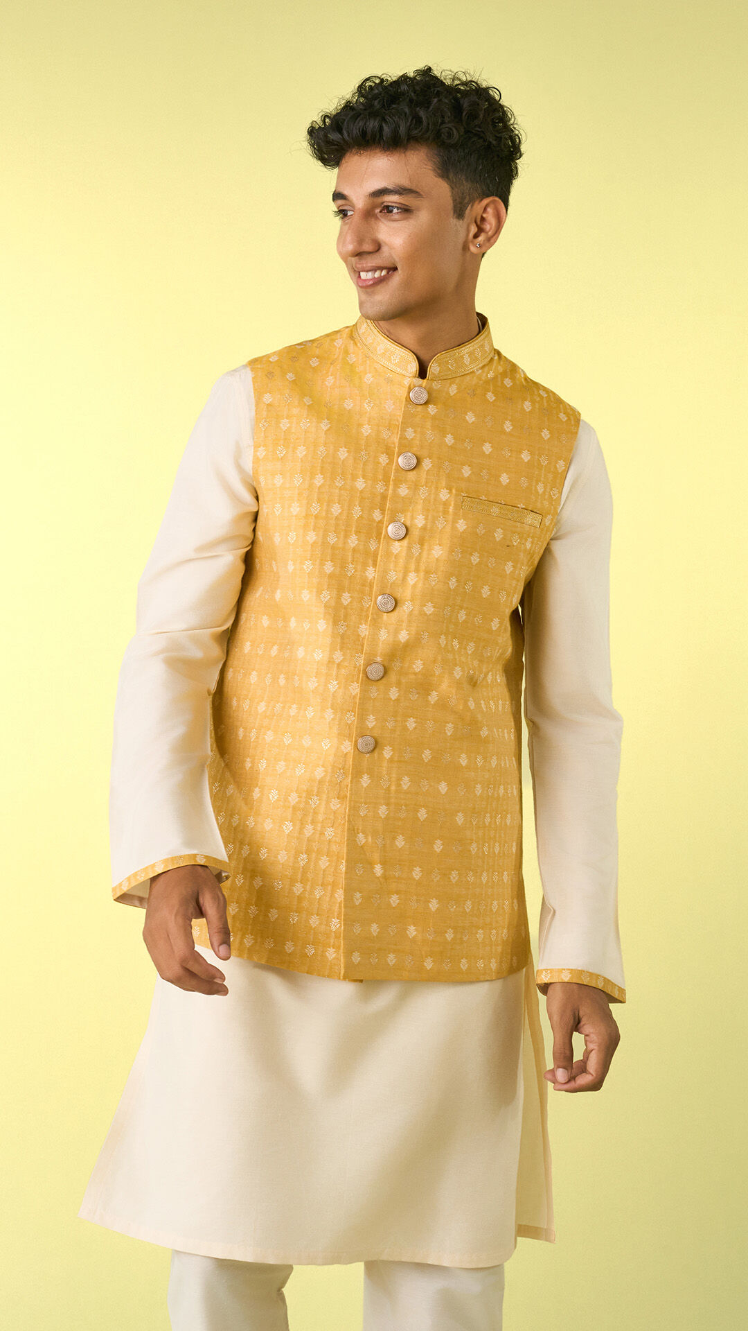 Young man wearing an ivory kurta paired with a mustard yellow embroidered Nehru jacket, featuring intricate woven motifs and a mandarin collar.